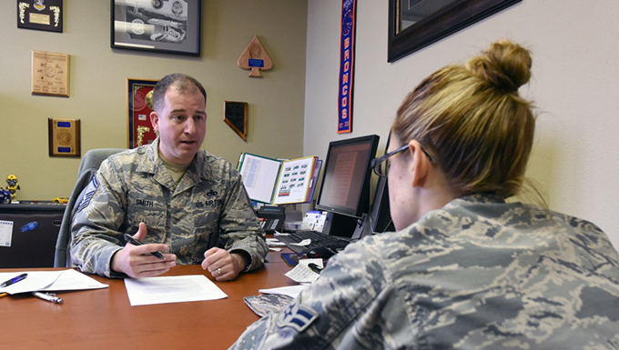 Two airmen speaking in an office at a table.